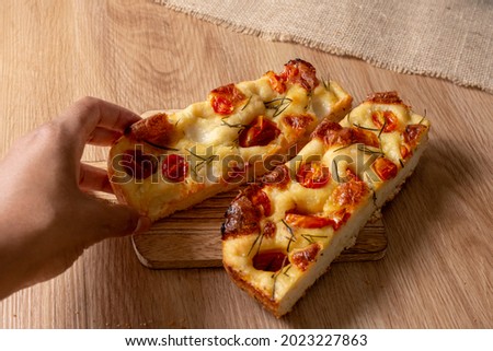 Hand holding slice of focaccia with tomato, rosemary, and olive oil