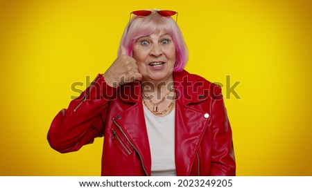 Elderly granny rocker woman with pink hair looking at camera doing phone gesture like says hey you call me back. Senior old grandmother emo in red jacket isolated on yellow background. People emotions