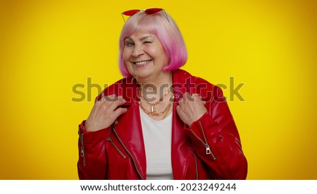 Playful happy elderly rocker granny woman in jacket blinking eye, looking at camera with toothy smile, winking and flirting, expressing optimism. Senior mature old emo grandmother on yellow background