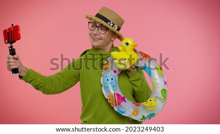 Mature grandmother traveler blogger with swimming ring and inflatable duck toy taking selfie on mobile phone make video call online. Studio shot. Senior woman on summer holiday vacation, trip. Tourism