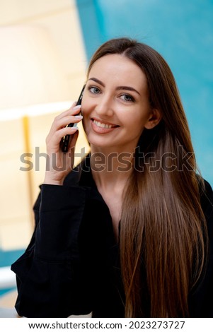 Cheerful young woman in casual style standing in modern room and talking by phone. Pretty woman have a phone call while smiling to the camera