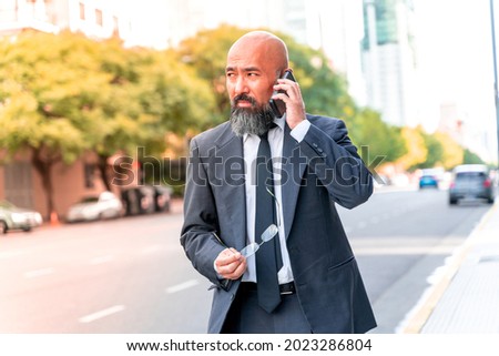 Serious Asian businessman talking on the phone standing on the street in an urban environment.
