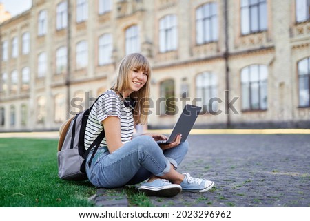Young cute caucasian blonde female student at university campus sitting on green grass with backpack and laptop browsing internet happy emotion looking straight at camera. High quality photo