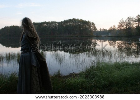 Cloaked Medieval woman walking by misty early morning lake. Green Witch or Shamanic story. Royalty-Free Stock Photo #2023303988