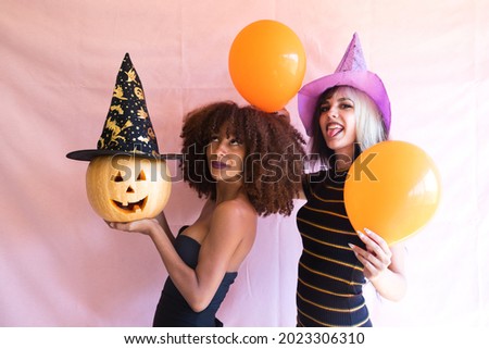 Two women, one blonde and the other Afro-American, young and beautiful, are having fun at a party playing with a Halloween pumpkin. They have confetti and streamers. Concept of happiness