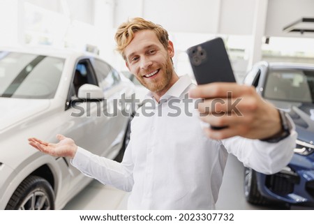 Man customer happy buyer client in white shirt do selfie shot on mobile cell phone show car choose auto want buy new automobile in showroom vehicle dealership store motor show indoor. Sales concept.