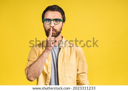 A serious handsome man keeps his front finger on his lips, tries to keep the conspiracy. Shh, please keep quiet. Attractive man showing a sign of silence isolated over yellow background.