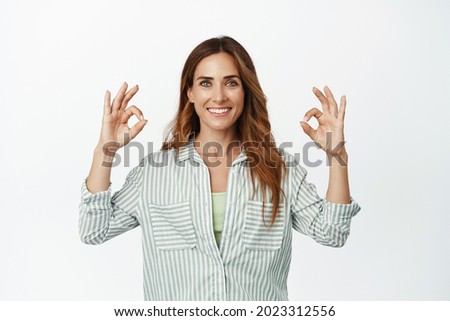 Portrait of confident businesswoman smiling happy, showing okay OK sign in approval, like and agree, recommend quality service, standing in blouse against white background