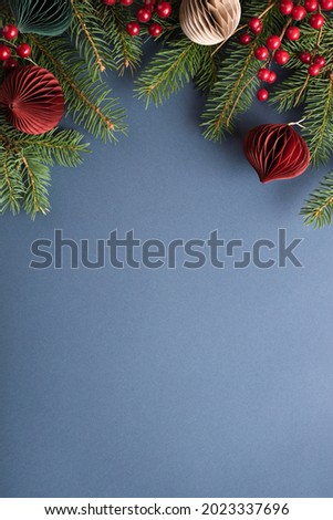 Christmas border with paper decorations, spruce branches and berries on blue background.