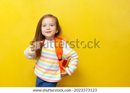 A cute beautiful girl with blue eyes and a backpack points her finger to the side. Yellow background, space for text. Back to school. Emotions, positive. September. High quality photo