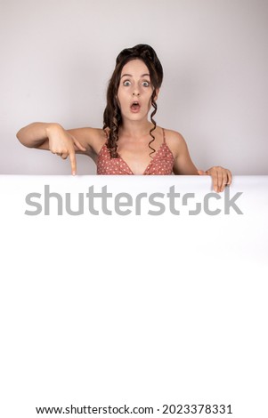 An attractive female slim model with long brown auburn hair on a white background wearing a vintage dress holding up a blank sign board or advertising banner pointing down to the advert