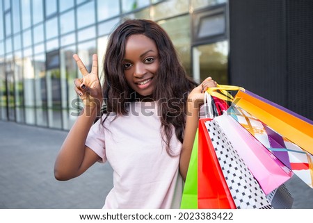 Peace Gesture Sign! Afro american lady hold many bags shopper african woman dressed plain t-shirt carrying enjoying new clothes packs things after shopping buyings sales black friday concept