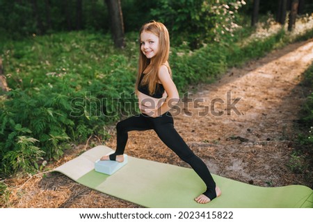 Hardworking, beautiful blonde preschool girl, child athlete performs an exercise, warm-up, stretching in gymnastics, yoga, standing on a green rug with a blue cube in the forest at sunset.