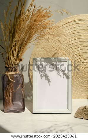 Modern still life with shadows and sunlight. White blank wooden frame mockup. Bouquet of dry herbs in the bottle, napkin and stone. Empty picture frame. Interior design concept. 