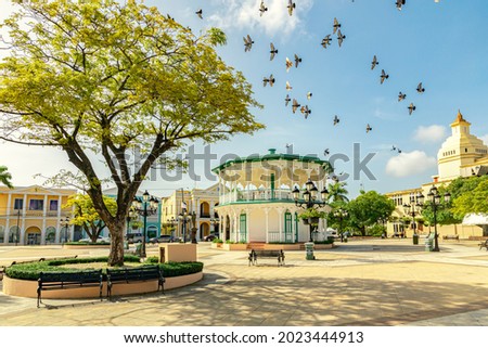 Flying birds in the sky of the Central Park in Puerto Plata, Independence Square, Plaza de Independencia, and a catholic church in the downtown  Royalty-Free Stock Photo #2023444913