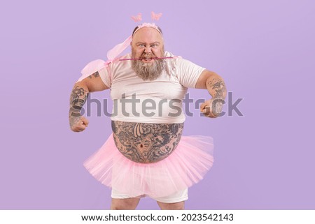Funny brutal obese man with different tattoos in fairy costume with wings holds magic stick in teeth on purple background in studio