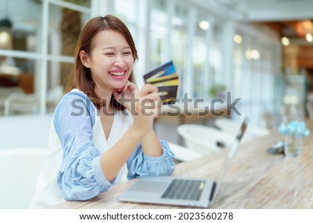 A beautiful Asian woman in a white shirt is sitting in front of a computer with a credit card in her hand to prepare for online shopping. Her face looks very happy in a bakery.