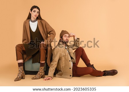 trendy woman sitting on vintage tv set near stylish man on floor on beige background Royalty-Free Stock Photo #2023605335