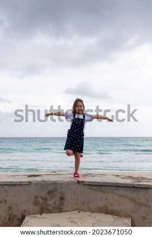 the child stands on one leg, arms outstretched to the sides against the background of the sea