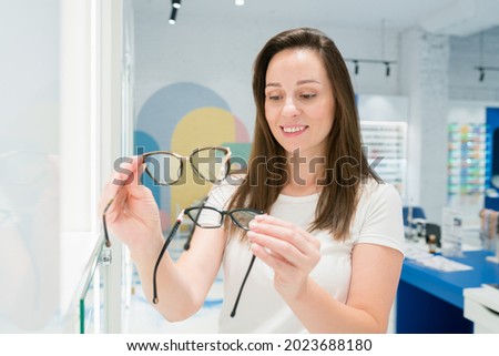 Vision and eye care concept. Cute caucasian woman choosing glasses in optical store. Female holding two frames and selecting  the perfect eyewear for her face shape  Royalty-Free Stock Photo #2023688180