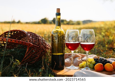 Picnic, romantic evening in nature concept. Bottle of red wine, glasses with drink, grapes, peaches on wooden board, wicker picnic basket on sunset background Royalty-Free Stock Photo #2023714565