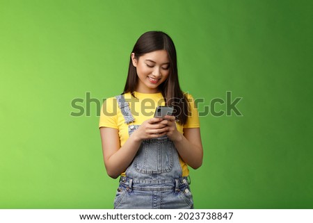 Outgoing cute asian brunette woman scroll online shop, purchase summer tour internet, hold smartphone, look amused telephone screen, use app, edit photos post social media, stand green background