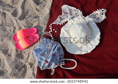Photo of a beach still life. Bright pink beach slippers sit on the sand, while a nautical-patterned bag, a beautiful ladies' beach hat with a lace ribbon and seashells lie on a burgundy beach blanket.