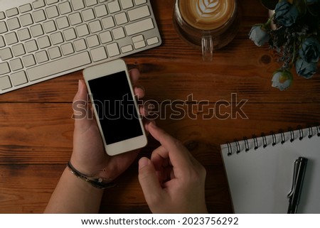 Above view and table top view with male hands using smartphone on the wooden table with modern keyboard, latte coffee and writing note.