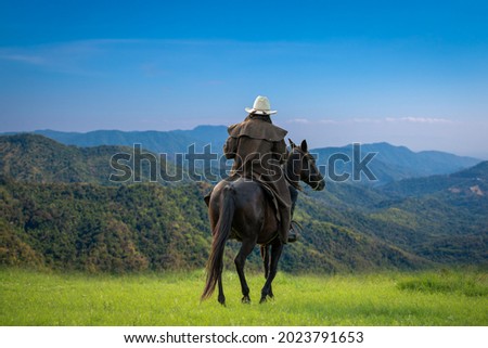 Back side view cowboy riding on horseback ,he wearing western dresser style and hat on summer with green mountain and blue sky background.  