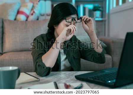 chinese lady sitting at table is getting fatigued eye from gazing at pc screen without resting. asian female removing her glasses and rubbing her eye to ease the pain. health and eye care concept Royalty-Free Stock Photo #2023797065