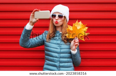 Portrait of stylish young woman taking selfie by phone with yellow maple leaves blowing her lips wearing a white hat, jacket on red background