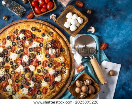 On a dark blue background, a large pizza on a wooden plate with many ingredients. Bright composition. High angle view. Restaurant and hotel business, pizzeria.