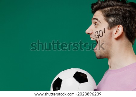 Side view european man fan supporter wears basic pink t-shirt cheer up support favorite football sport team hold in hand soccer ball watch tv live stream isolated on dark green color background studio