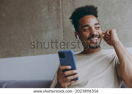 Young fun smiling african american man in beige t-shirt sitting on grey sofa indoors apartment use air pods mobile cel phone listen to music podcast looking aside resting on weekends staying at home
