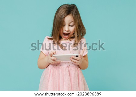 Fun cute kid girl 5-6 years old wears pink dress use play app on mobile cell phone hold gadget for pc video games isolated on pastel blue color background child studio Mother's Day love family concept