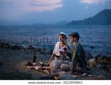 a young man and a woman drink red wine on the beach from glasses, sitting on a blanket in the evening, they have a camera, take pictures of the landscape.