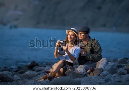 a young man and a woman drink red wine on the beach from glasses, sitting on a blanket in the evening, they have a camera, take pictures of the landscape.