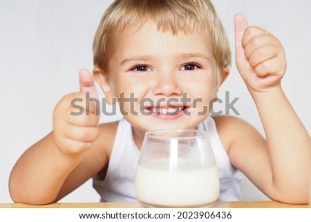 a smiling blond boy with brown eyes drinks milk from a glass at a wooden table and shows his class with his hands.

