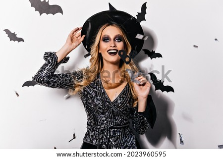 Excited glamorous witch with black makeup laughing to camera. Smiling blonde vampire in hat relaxing in halloween.