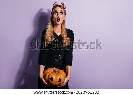 Shocked long-haired woman posing after halloween masquerade. Studio photo of beautiful blonde girl with pumpkin.