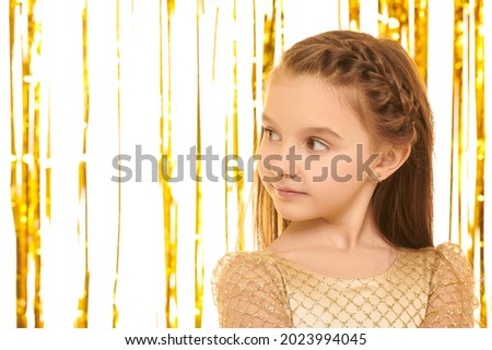 Close up portrait of beautiful child girl in a golden evening dress looking away on a background with shiny gold foil curtains. Children's party. Kid's fashion and holidays. 