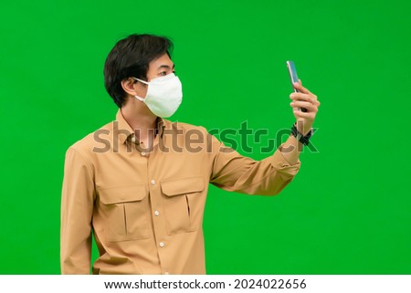 A half-body of an Asian man in a yellow shirt holding his phone wearing a medical mask stands behind a green screen.