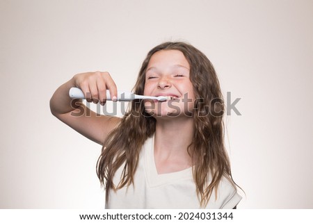 A portrait of a teenage Caucasian girl smiling, brushing her teeth, eyes closed, holding a tooth brush, isolated on the beige background