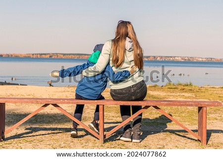 Mom and son sitting on bench by lake shore and pretend to be flying at sunny autumn day. Back view. Lifestyle, mothers tenderness. Son's day or mother's day concept