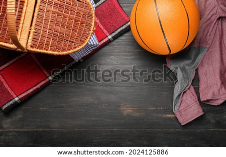 Picnic basket with plaid, ball and clothes on dark wooden background