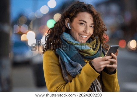 Young beautiful woman using mobile phone at dusk in the city while listening music through earphones. Happy woman using smartphone to do a video call on a winter day while looking at camera. Royalty-Free Stock Photo #2024209250