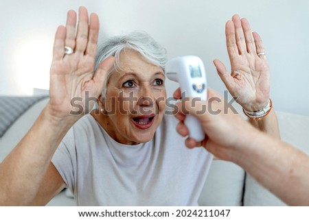 Senior woman looks straight ahead with arms raised, while an unrecognizable healthcare professional takes her temperature with an infrared thermometer at home. Senior woman getting temperature checked
