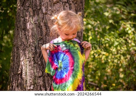 A little girl looking at her tie dye tee shirt- A little girl holding her new shirt up to her and looking at the tie dye design on it- A child is standing outside by a tree admiring her new shirt 