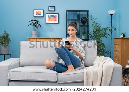 Contented woman sits on the sofa in the living room holding her phone in her hands, a girl sends messages to friends, writes a blog post, replies to emails, chats, taps on the smartphone screen