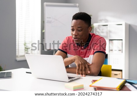 Trainee in office sits at desk in front of laptop monitor in background whiteboard, young dark-skinned man talks online with clients, co-workers, uses webcam, video call Royalty-Free Stock Photo #2024274317